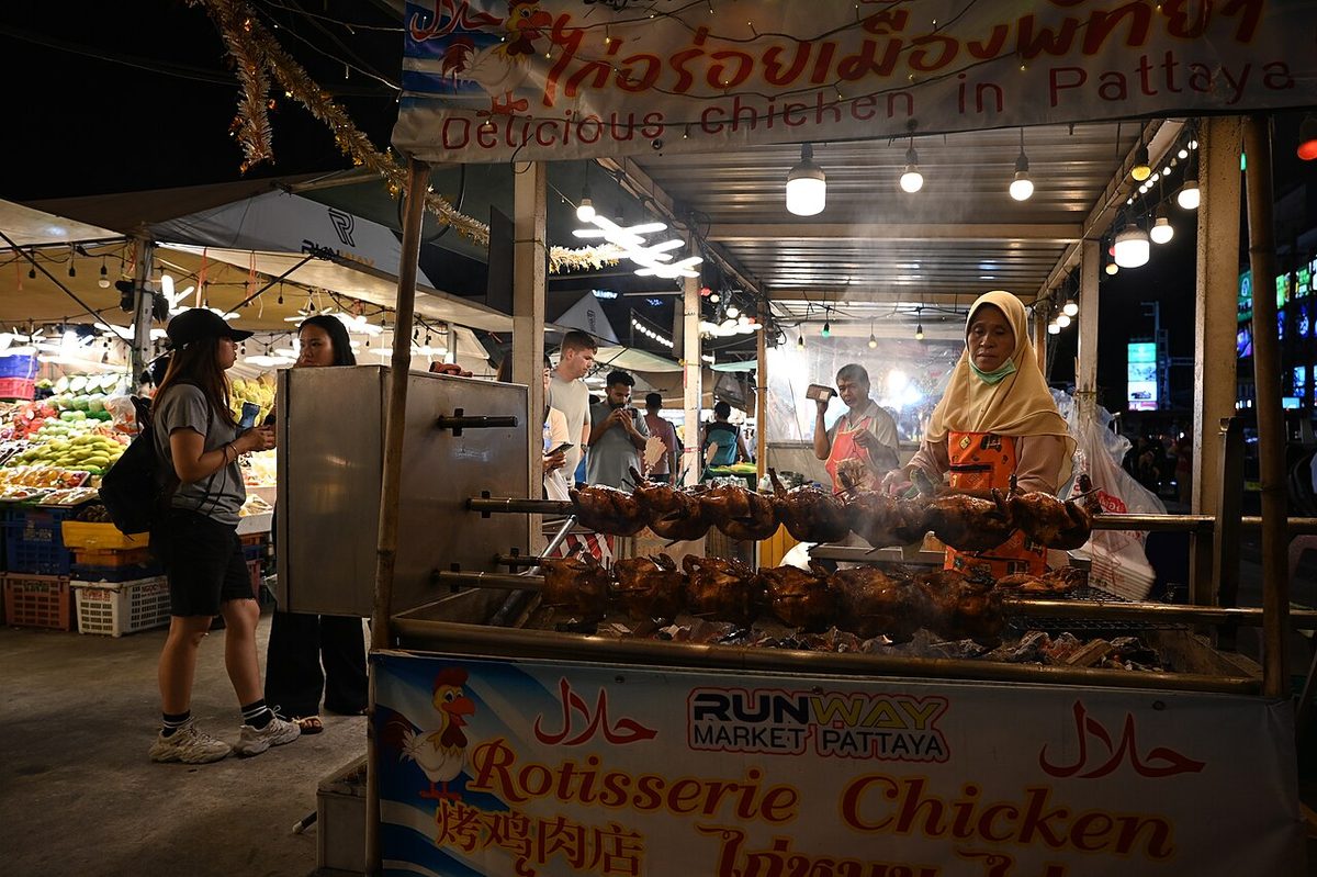 DFC 4674 Night market scene in Pattaya a vendor tends her rotisserie chicken stall while visitors browse nearby produce